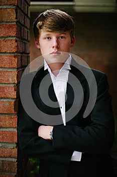 A blond teen in a tuxedo leaning on a brick wall.
