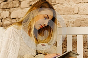 The blond teenage girl is holding the tablet and Teddy bear. Close-up view.