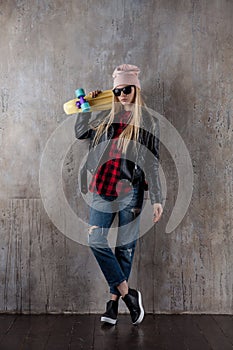 Blond teenager girl in black sunglasses and pink hat with blue skateboard. In front of concrete wall.