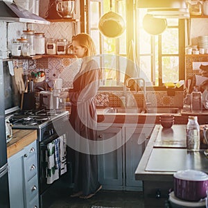 Blond tired woman making coffee in an old narrow cluttered kitchen at sunset