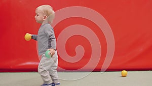 Blond toddler holds two little balls in hands and goes to his mother and then walks away