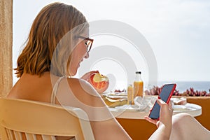 Blond Vegetarian Caucasian woman sit on a chair in the terrace of a hotel, eating and apple and checking the mail in her smart