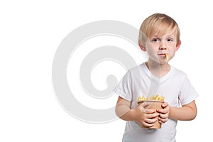 A blond, white-skinned child holds a disposable cup of French fries and a slice of fast food sticks out of his mouth. Happy boy in