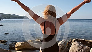 Blond woman arms outstretched on the beach at sunrise. Happy girl enjoying life in a black swimsuit early in the morning