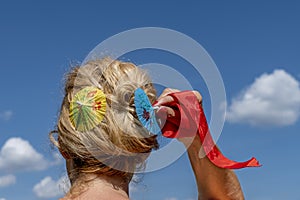 A blond woman arranges her hair using two colored paper umbrellas and a red ribbon, against the blue sky