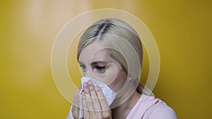Blond woman blowing her nose using white paper tissue against yellow background, flu and illness concept