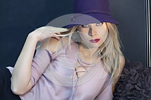 A blond woman with blue eyes and a purple hat and lilac laced blouse.