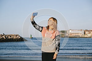 blond woman with casual clothes uses the mobile phone on the beach of a city at sunset with sunny day