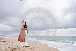 Blond woman Christmas tree sea. Christmas portrait of a happy woman walking along the beach and holding a Christmas tree