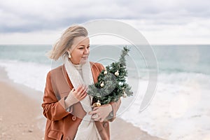 Blond woman Christmas tree sea. Christmas portrait of a happy woman walking along the beach and holding a Christmas tree