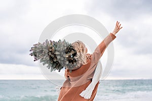 Blond woman Christmas tree sea. Christmas portrait of a happy woman walking along the beach and holding a Christmas tree