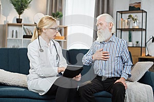 Blond woman doctor in glasses and white medical coat, visits her senior patient at home. Therapist is taking notes on