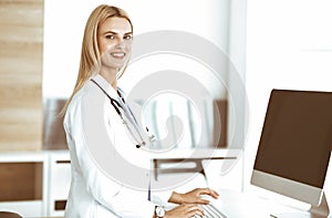Blond woman-doctor is typing on desktop computer at reception desk in clinic. Physician is excited and happy of her