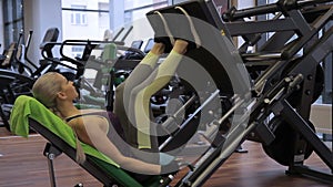 The blond woman is doing leg presses in the bright gym with modern equipment.