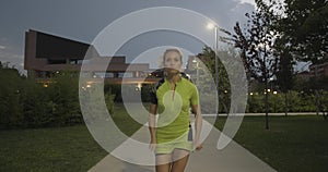 Blond woman in fitness wear running training on park walkway.Front following view.Summer evening or night.Industrial