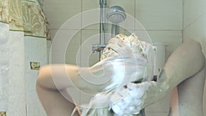 Blond woman foaming shampoo on hair in bathroom, water falling down from shower head, washing hair and body care