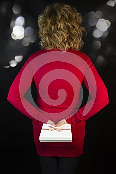 A blonde woman hides a gift behind her back against a black background with bokeh of lights