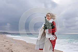 Blond woman holding Christmas tree by the sea. Christmas portrait of a happy woman walking along the beach and holding a