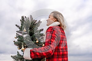 Blond woman holding Christmas tree by the sea. Christmas portrait of a happy woman walking along the beach and holding a