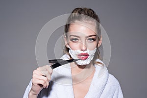 Blond woman with impudent look wearing white bathrobe isolated on gray background. Morning shaving, young female with