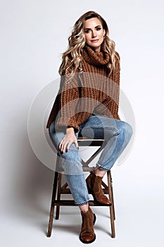 Blond woman with long curly hair sitting on chair dressed in comfy oversize knitted brown sweater