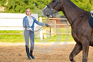 Blond woman with long hair jockey rider on a bay horse, in a paddock on a ranch