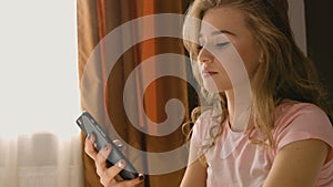 Blond woman sitting on the bed, holding her phone in hands and chatting to hear friends in a pink pajama.