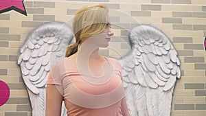 A blond woman stands on the background of a wall with bas-reliefs in the form of angel wings.