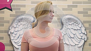 A blond woman stands on the background of a wall with bas-reliefs in the form of angel wings.