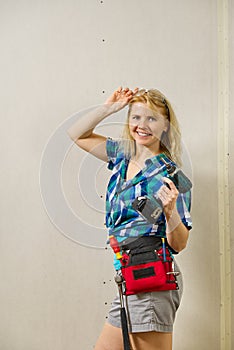 Blond woman wearing a DIY tool belt full of a variety of tools on a unpainted plasterboard wall background. Construction woman