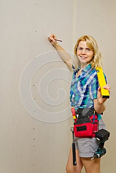 Blond woman wearing a DIY tool belt full of a variety of tools on a unpainted plasterboard wall background. Construction woman