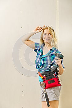 Blond woman wearing a DIY tool belt full of a variety of tools on a unpainted plasterboard wall background. Construction woman