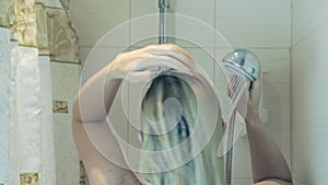 Blond woman wets her hair with water falling down from shower head in bathroom, body care concept