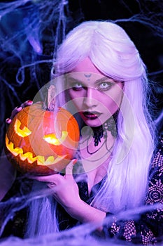 Blond woman witch with long white hair holding pumpkin. White lenses in the eyes. Halloween party.