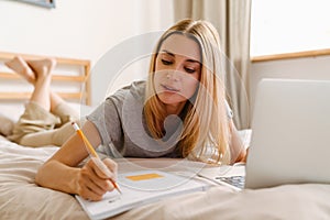 Blond woman writing down notes and using laptop while lying in bed