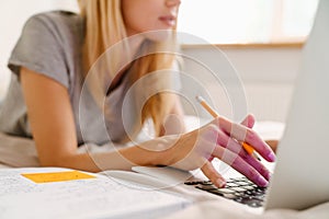 Blond woman writing down notes and using laptop while lying in bed