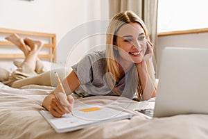 Blond woman writing down notes and using laptop while lying in bed