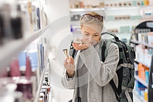 Blond young female traveler wearing travel backpack choosing perfume in airport duty free store.