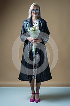 Blond young smiling happy woman in coat and dress with bouquet  on beige background studio