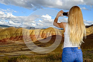 Blond young woman taking cell phone photos of the Painted Hills Unit - John Day Fossil Beds National Monument, Oregon, USA
