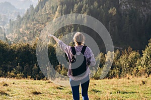 Blonde 30s girl in sunglasses pointing finger on mountains background. Young female with backpack hiking in nature