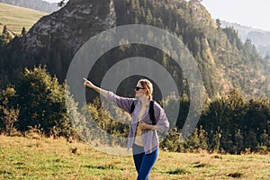 Blonde 30s girl in sunglasses pointing finger on mountains background. Young female with backpack hiking in nature