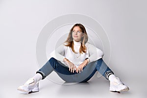Blonde adolescent in jeans, sweater, socks and sneakers. Smiling, sitting on floor, legs wide apart, isolated on white. Close up