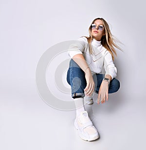 Blonde adolescent in sunglasses, watch, jeans, sweater, socks and sneakers. She squatting on floor, isolated on white. Close up