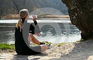 Blonde athlete girl sitting on ground to relax after jogging drinking water under a tree on a lake shore