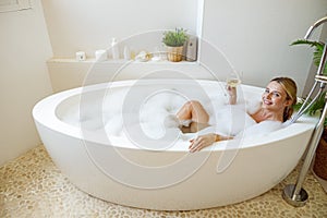 Blonde beautiful young woman lying in bathtub with foam and holding glass of white wine.