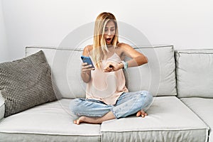 Blonde beautiful young woman sitting on the sofa at home using smartphone looking at the watch time worried, afraid of getting