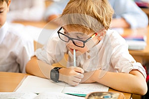 Blonde boy with big black glasses sitting in classroom, studing, smiling. Education on elementary school, first day at school