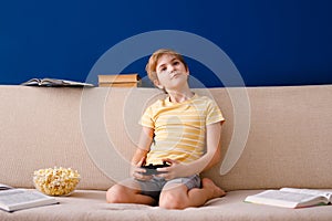 Blonde boy plays with gamepad instead of lessons and eats popcorn