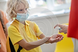 Blonde boy in preventive mask disinfecting his hands while enetering the school
